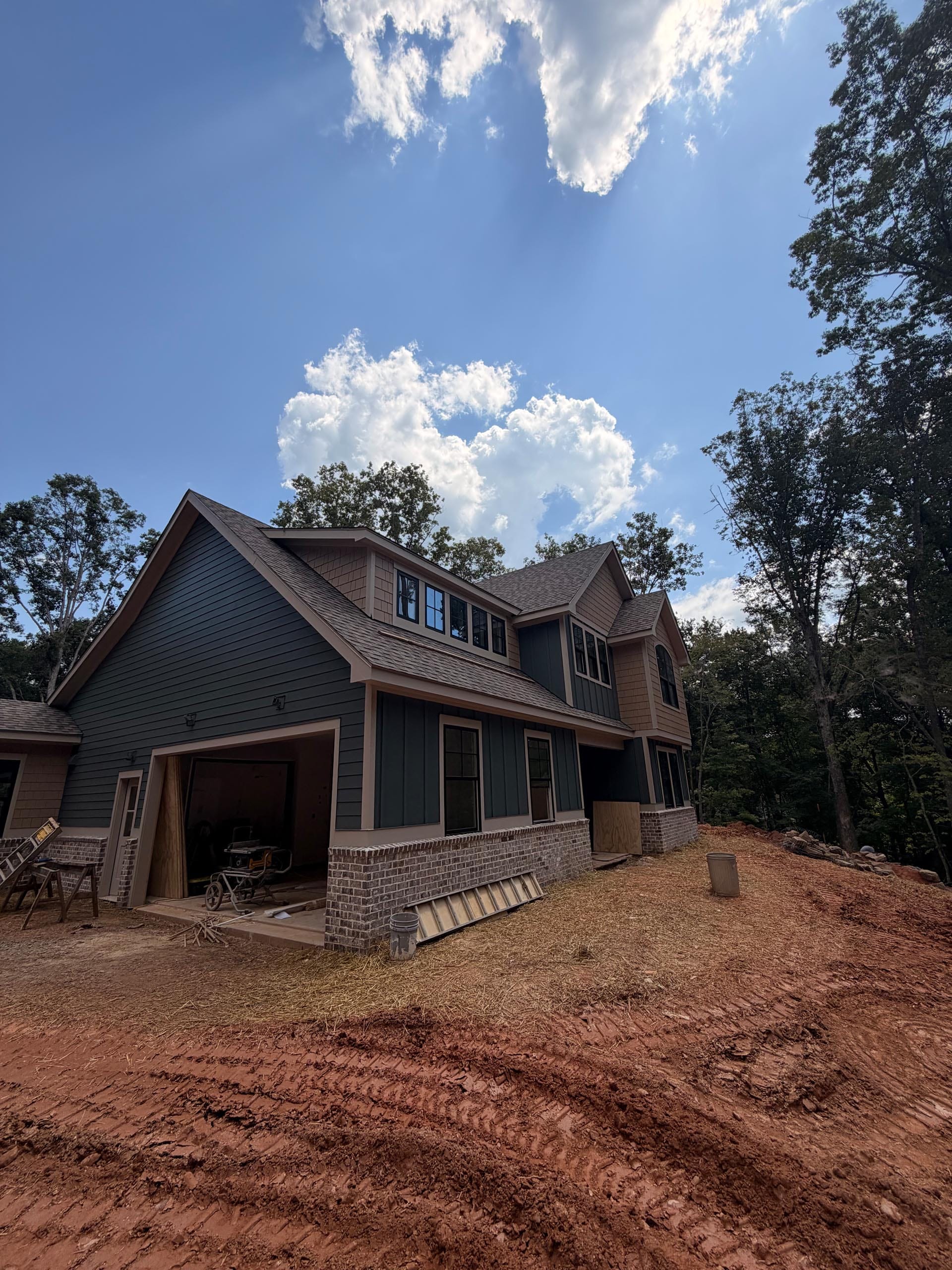 A two-story house under construction with a brick and green exterior, open garage, and dirt yard, surrounded by trees under a partly cloudy sky.