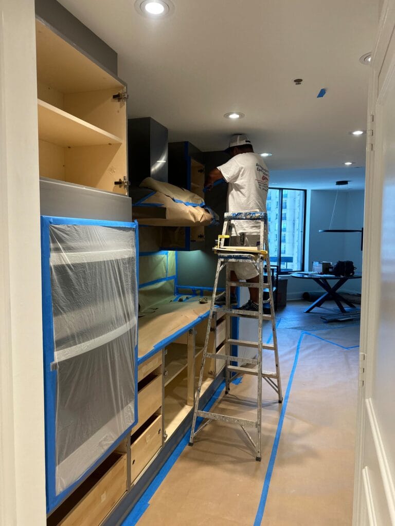 A worker on a ladder paints or repairs upper kitchen cabinets in a modern apartment under renovation, with floors and surfaces covered for protection.