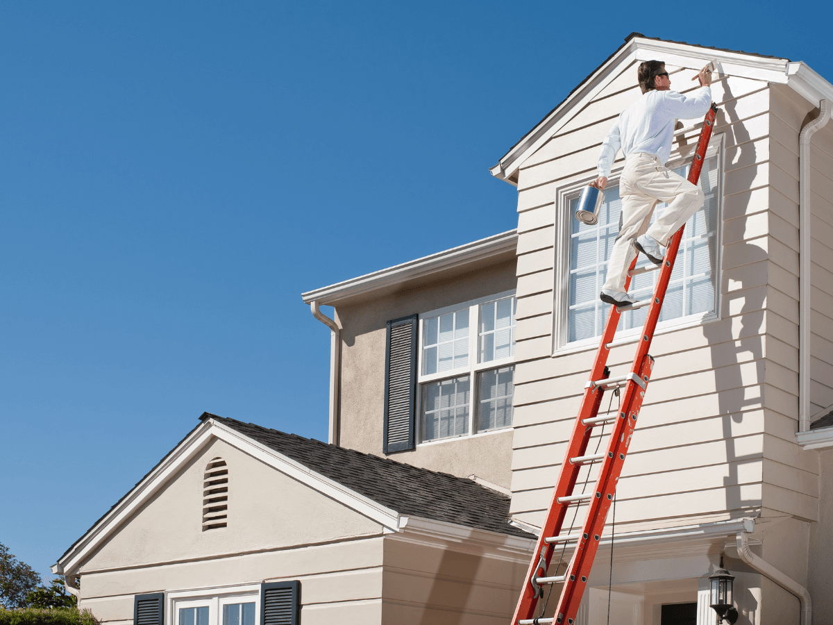 A person stands on a red ladder, engaged in seasonal painting, carefully painting the trim near the roof of a beige house with white and black accents on a clear, sunny day.